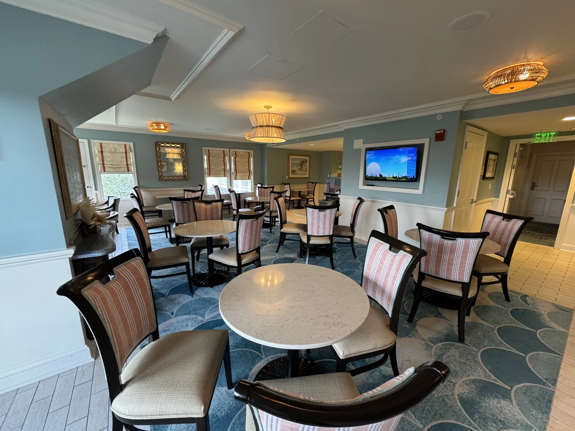 Un salon bien éclairé avec des tables rondes, des chaises rembourrées à rayures, des murs bleus, des œuvres d'art et une télévision murale - parfait pour se détendre ou recevoir l'assistance d'un concierge Aux Invités inspiré par l'hospitalité de Walt Disney World.