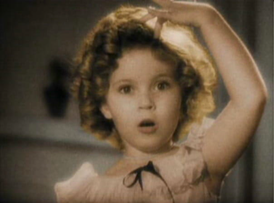 Une petite fille aux cheveux bouclés, vêtue d’une robe claire, porte une main à sa tête et regarde devant elle avec un air surpris — comme un enfant qui reliait deux mondes, le 3 mars 1937.