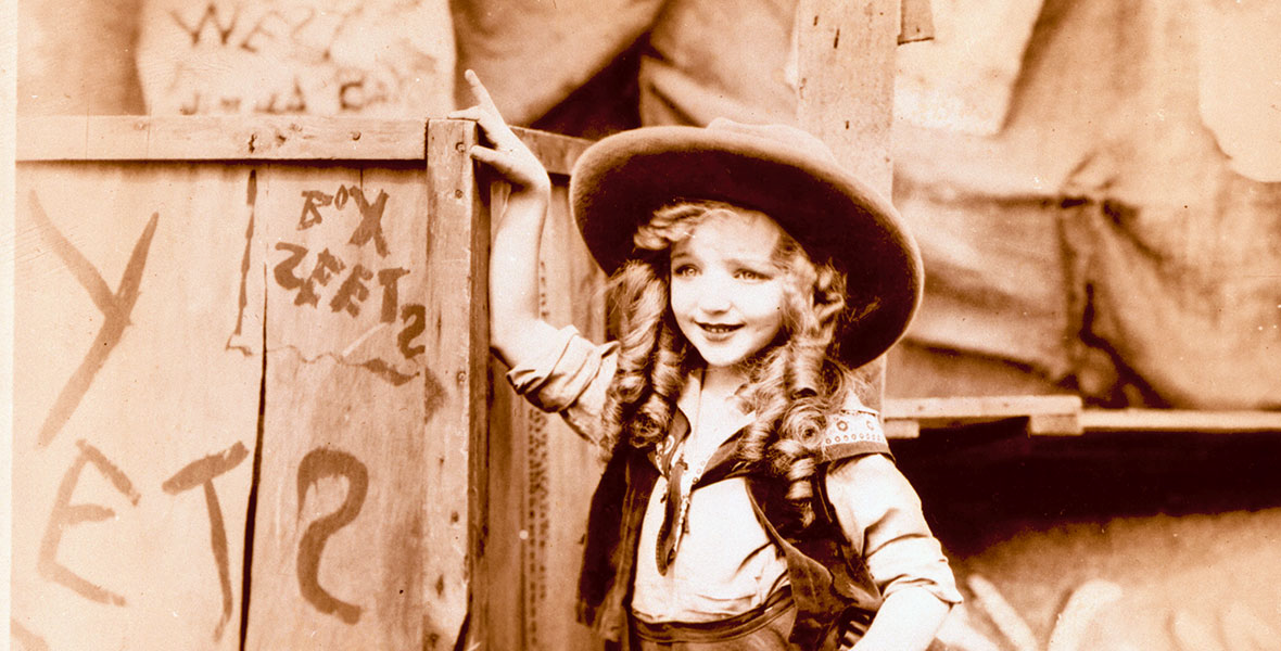 Photo aux tons sépia d'un jeune garçon, qui rappelle Bobby Driscoll, coiffé d'un chapeau de cow-boy et vêtu d'un costume western, souriant et adossé à une clôture en bois couverte de graffitis.