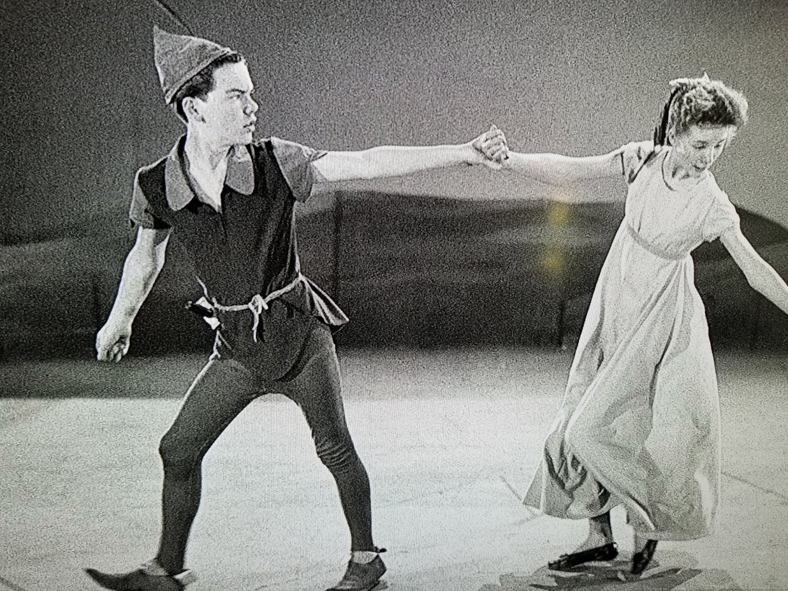 Deux artistes sur scène : l'un, déguisé en Peter Pan et rappelant Bobby Driscoll, tient la main d'une petite fille en robe et la guide doucement vers l'avant.