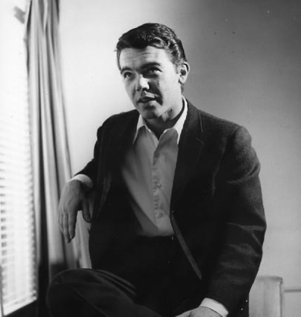 Un homme vêtu d'une veste de costume et d'une chemise au col ouvert est assis à l'intérieur près d'une fenêtre équipée de stores et de rideaux, le regard légèrement levé. Cette image en noir et blanc dégage une atmosphère intemporelle, qui rappelle les portraits classiques tels que ceux de Bobby Driscoll.