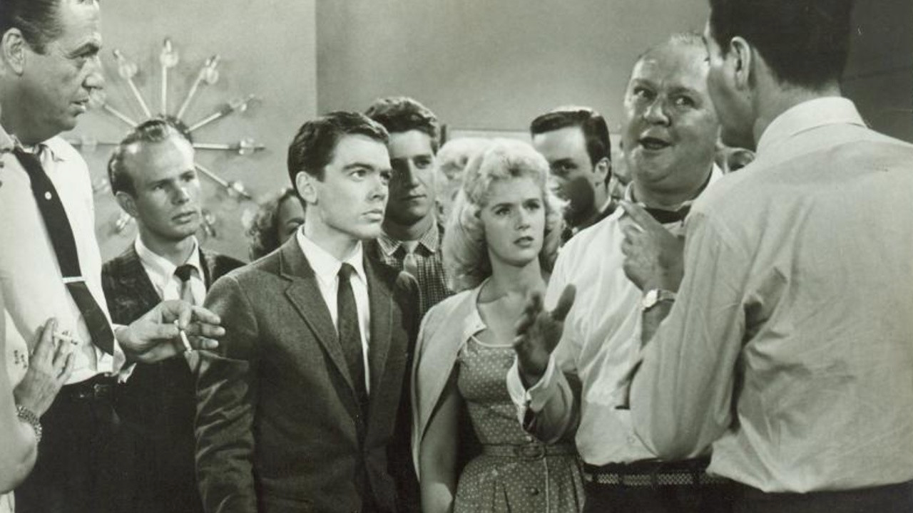 Un groupe de personnes vêtues à la mode des années 1950 se tient à l'intérieur, plongé dans une discussion sérieuse, tandis que certaines regardent un homme — qui rappelle peut-être Bobby Driscoll dans ses jeunes années — en train de parler au premier plan.