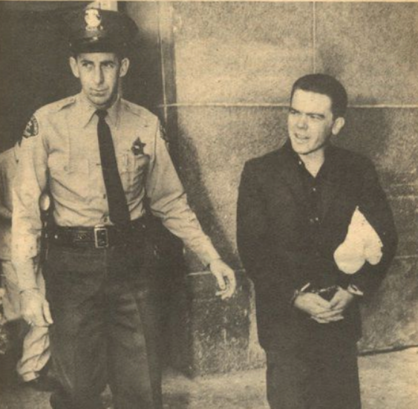 Un policier en uniforme escorte un homme menotté tenant un tissu blanc ; ils marchent ensemble devant un bâtiment en pierre — une scène qui rappelle l'époque du 3 mars 1937 et des récits tels que « Bobby Driscoll, l'enfant qui reliait deux mondes ».