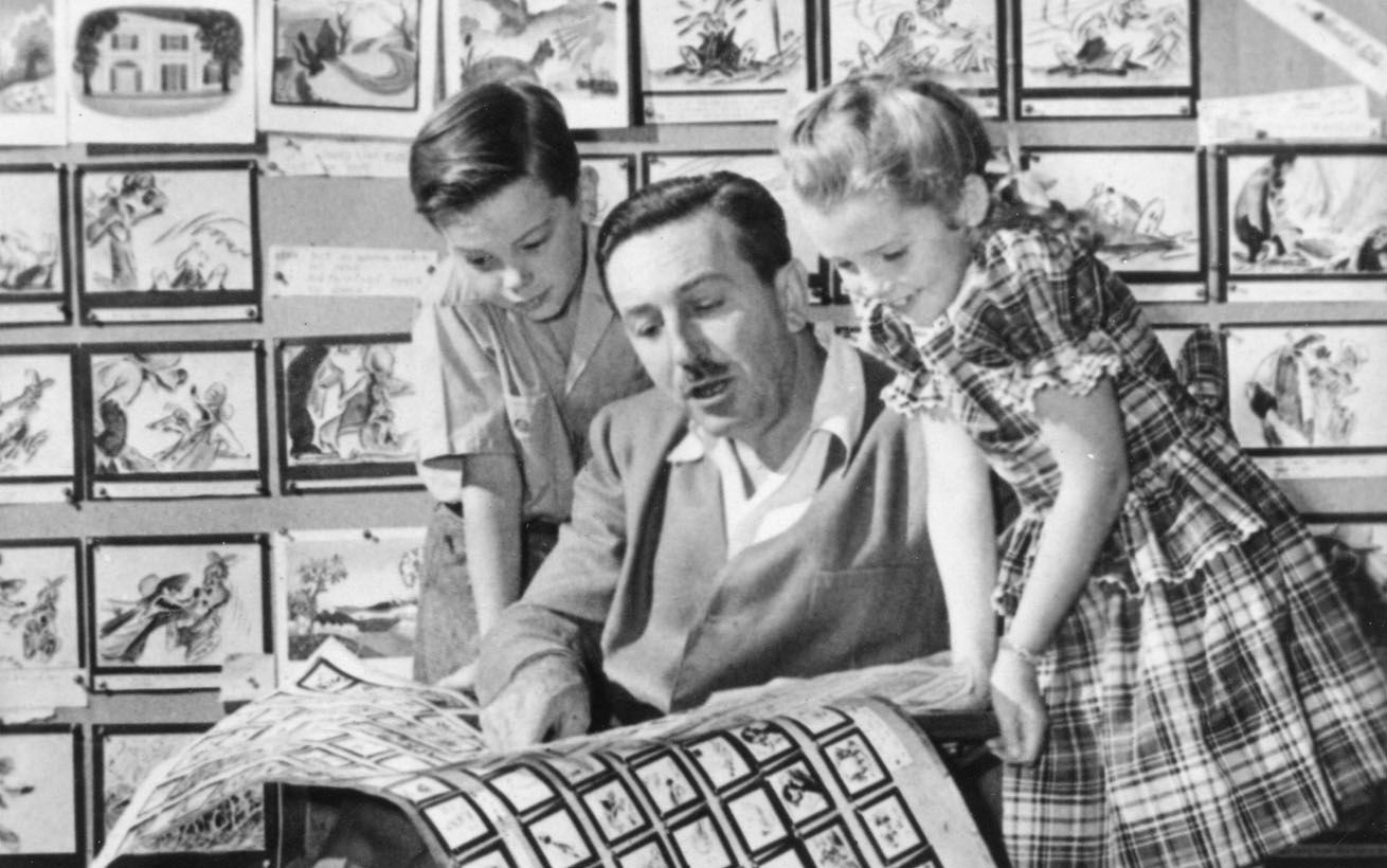 Un homme montre à deux enfants un grand storyboard dont les cases illustrées recouvrent le mur derrière eux, évoquant des scènes du film « 3 mars 1937 » et rappelant l’émerveillement juvénile de Bobby Driscoll dans son enfance.