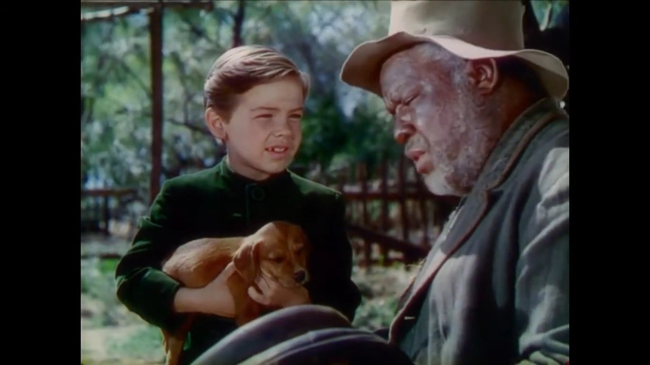 Un jeune enfant tenant un petit chien brun se tient à côté d'un homme plus âgé coiffé d'un chapeau, en plein air, avec des arbres et une clôture en bois en arrière-plan — une scène qui rappelle Bobby Driscoll, le 3 mars 1937.