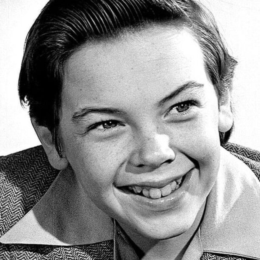 Photo en noir et blanc en gros plan de Bobby Driscoll, un jeune garçon souriant aux cheveux courts et soigneusement peignés, vêtu d'une chemise à col et d'une veste à motifs. Né le 3 mars 1937, son charme juvénile transparaît dans ce portrait.