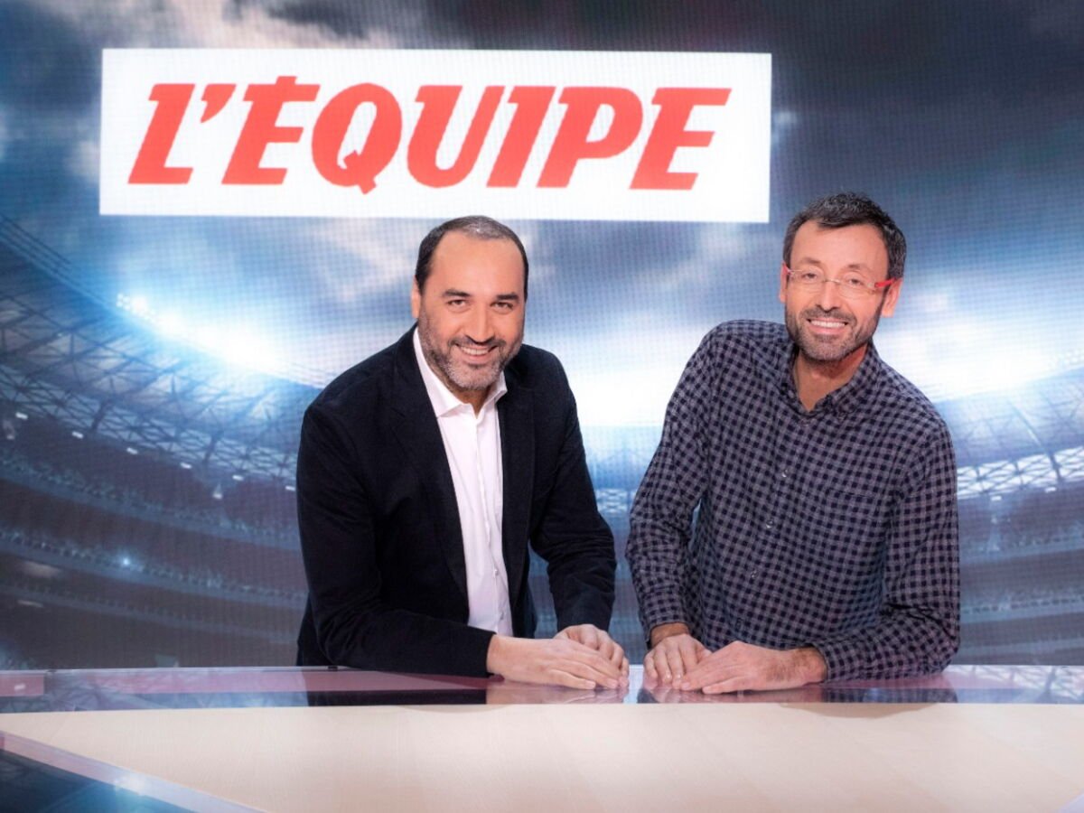 Deux hommes se tiennent derrière une table de rédaction devant un grand panneau "L'ÉQUIPE" sur fond de stade, représentant La Chaîne L'Équipe, la chaîne de sport gratuite.