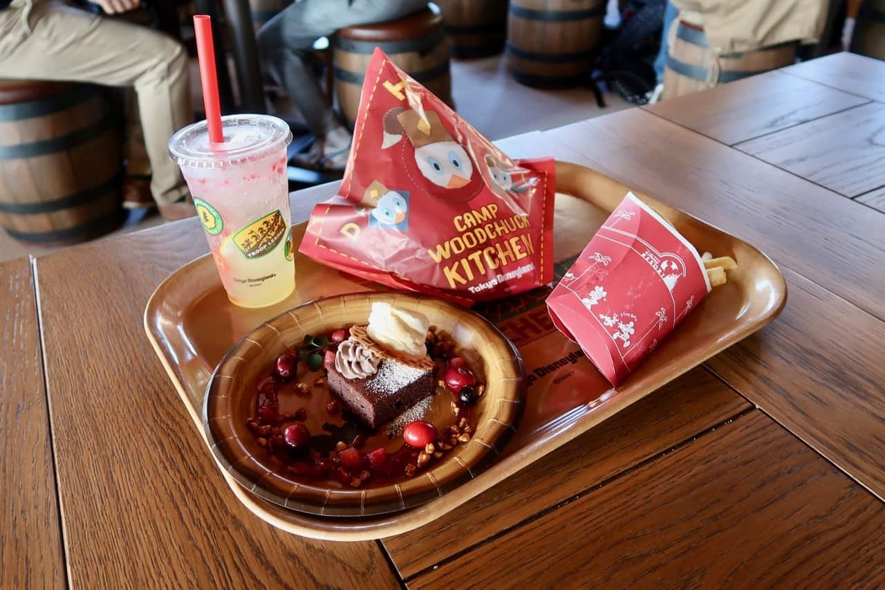 Un plateau avec une boisson, un dessert garni de canneberges et un biscuit, des frites et des snacks étiquetés "Camp Woodchuck Kitchen" sur une table en bois - parfait pour recueillir des avis sur les restaurants à Tokyo Disneyland.