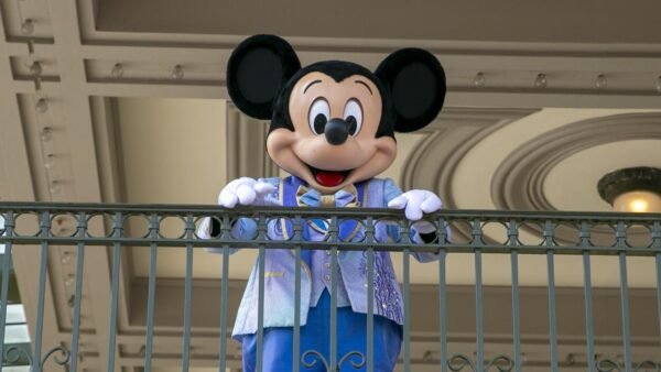 Une personne déguisée en Mickey Mouse se tient derrière une balustrade décorative, vêtue d'une veste bleu clair et d'un nœud papillon, capturant parfaitement l'image de marque Disney alors qu'elle sourit à l'appareil photo.