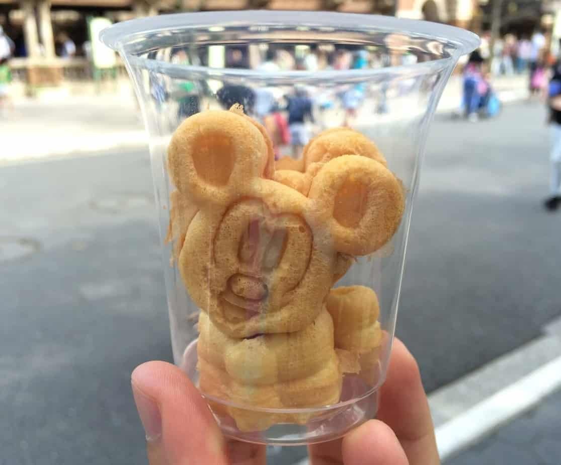 Un gobelet en plastique transparent contenant de petites pâtisseries en forme de tête de souris de dessin animé, présenté en extérieur avec des personnes et des bâtiments flous en arrière-plan - parfait pour une comparaison des Kiosques À Snacks Tokyo Disneyland et autres parcs à thèmes.