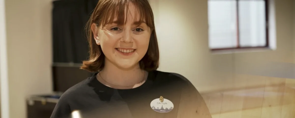Une femme aux cheveux et à la frange bruns, vêtue d'une chemise noire et d'un badge, sourit à l'appareil photo dans un décor intérieur, incarnant l'esprit amical des Cast Members de Disneyland Paris.