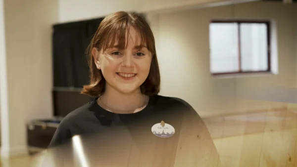 Une femme aux cheveux et à la frange bruns, vêtue d'une chemise noire et d'un badge, sourit à l'appareil photo dans un décor intérieur, incarnant l'esprit amical des Cast Members de Disneyland Paris.