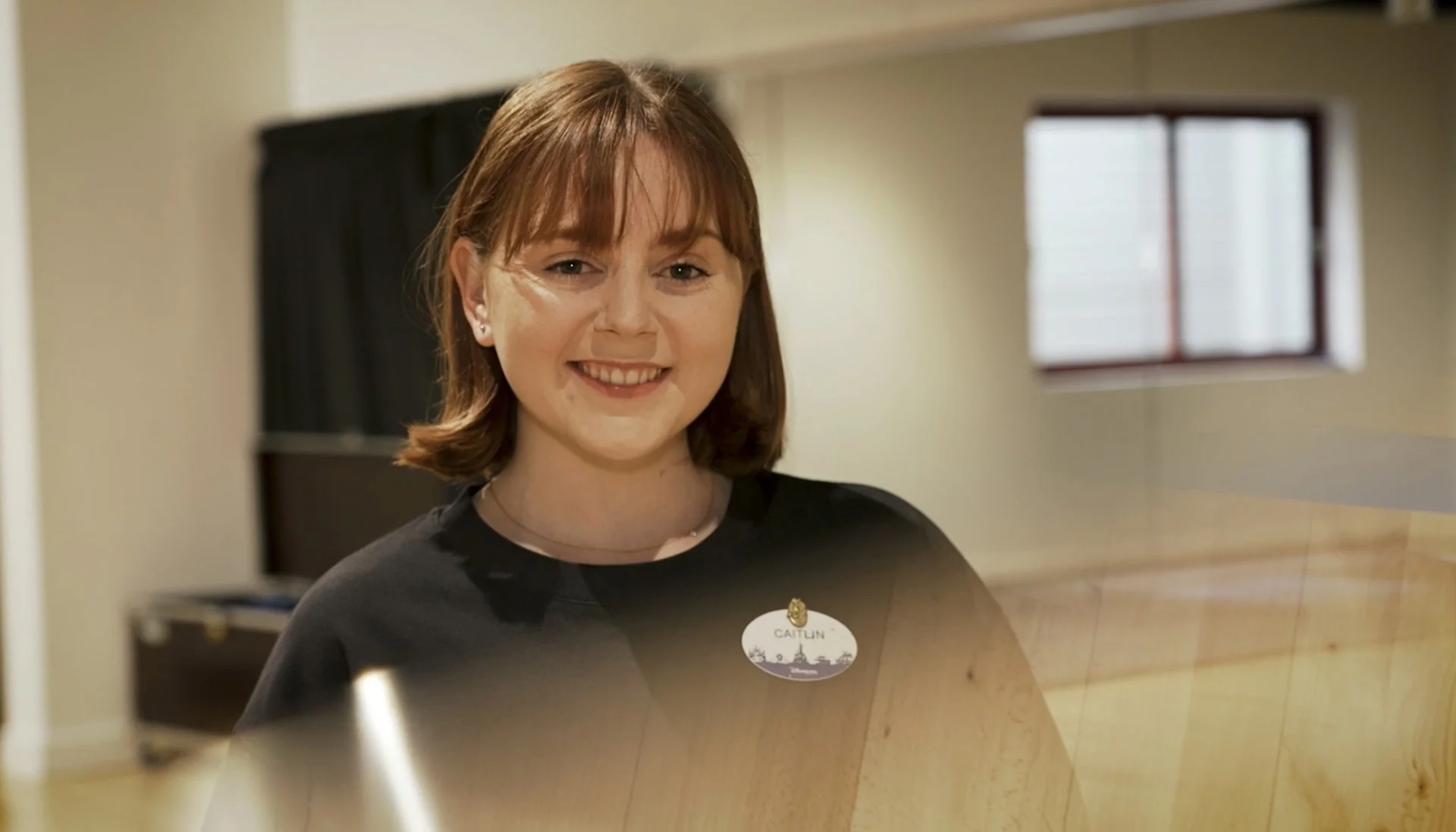 Une femme aux cheveux et à la frange bruns, vêtue d'une chemise noire et d'un badge, sourit à l'appareil photo dans un décor intérieur, incarnant l'esprit amical des Cast Members de Disneyland Paris.