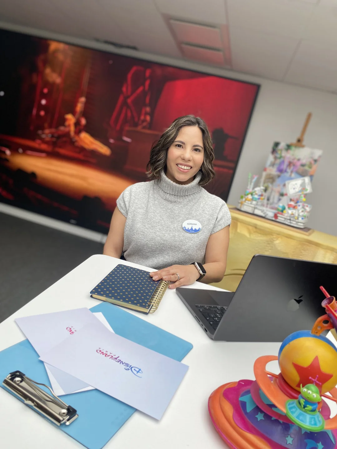 Une femme est assise à un bureau avec un ordinateur portable, un carnet de notes, des enveloppes et des jouets colorés, souriant à la caméra dans un bureau avec des fournitures artistiques et un grand écran - embrassant la Magie Disney qui inspire les Cast Members tous les jours.
