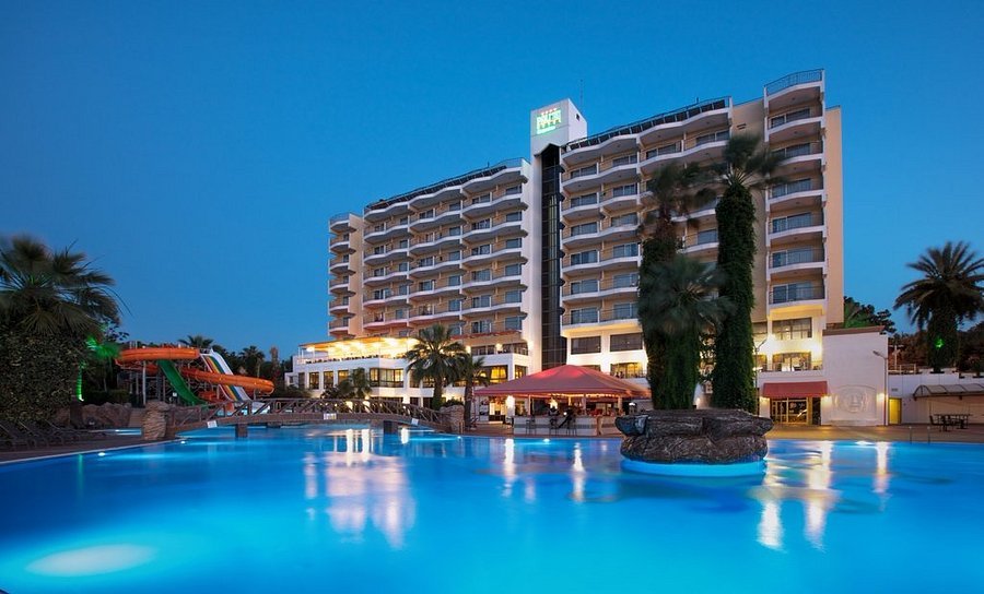 Au crépuscule, on aperçoit un grand hôtel 5 étoiles doté de balcons, donnant sur une piscine extérieure illuminée, entourée de palmiers et de toboggans aquatiques, proposant des chambres et des villas luxueuses à quelques minutes seulement de Disneyland Paris.