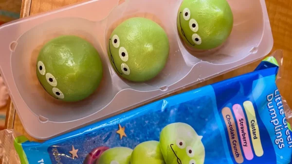 Trois boulettes de mochi vertes avec trois yeux chacune sont disposées dans un plateau en plastique à côté de leur emballage illustré "Little Green Dumplings", un en-cas emblématique des kiosques à snacks des célèbres parcs à thèmes de Tokyo Disneyland.