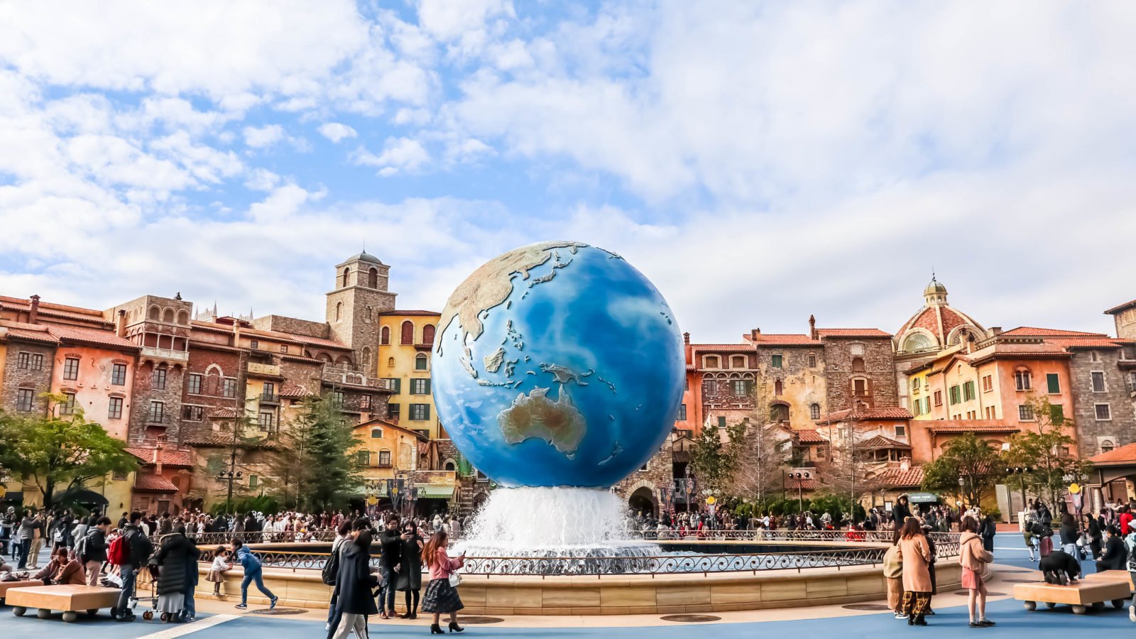 Une grande fontaine en forme de globe, pièce maîtresse d'une place, entourée de visiteurs, avec en arrière-plan des bâtiments colorés de style européen : l'une des attractions phares de Tokyo Disney.