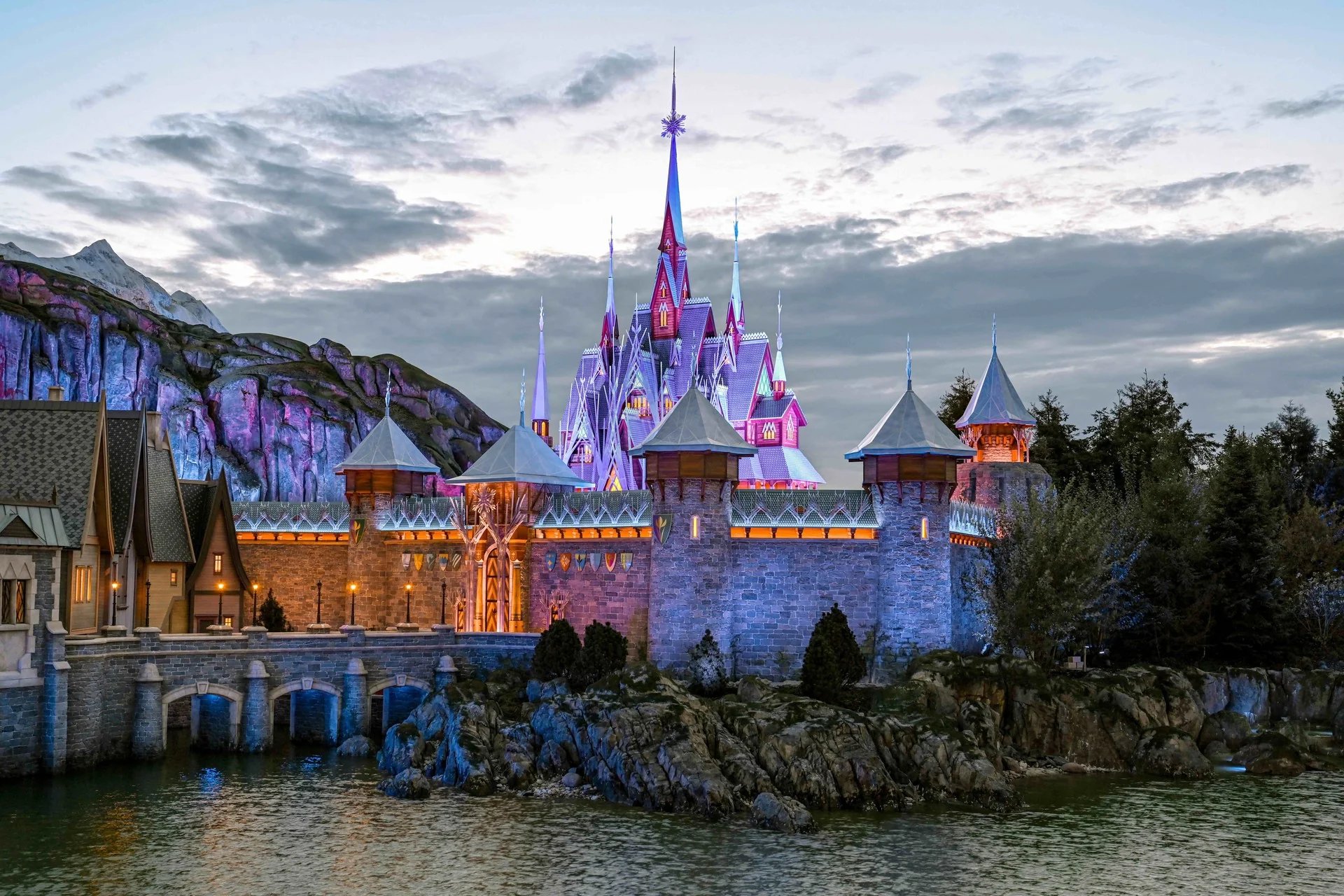 Un château lumineux inspiré du Château d'Arendelle, avec ses tours pointues et ses murs de pierre, se dresse au bord d'une étendue d'eau, au milieu d'un terrain rocailleux et de montagnes, sous un ciel nuageux.
