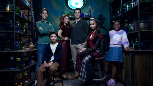 Cinq adolescents posent dans une pièce colorée et faiblement éclairée, remplie de bouteilles et d'étagères ; debout ou assis, ils sont tous tournés vers l'appareil photo, ce qui rappelle une scène de « Waverly Place » dans la saga Des Russo.