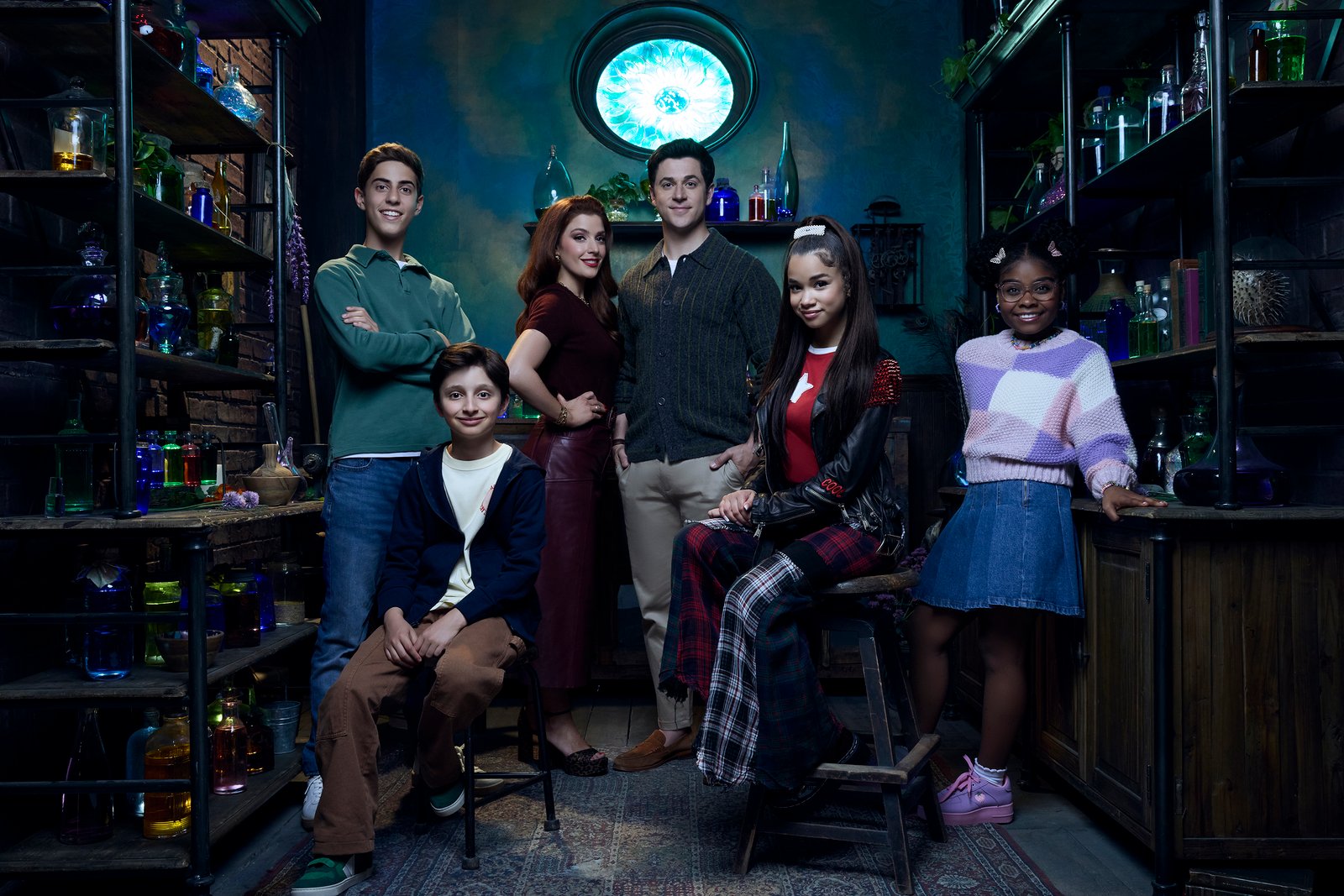 Cinq adolescents posent dans une pièce colorée et faiblement éclairée, remplie de bouteilles et d'étagères ; debout ou assis, ils sont tous tournés vers l'appareil photo, ce qui rappelle une scène de « Waverly Place » dans la saga Des Russo.