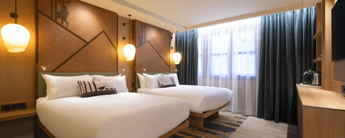 Découvrez le confort des chambres rénovées du Disney Sequoia Lodge : une chambre d'hôtel spacieuse dotée de deux lits doubles, d'œuvres d'art accrochées aux murs, de suspensions élégantes, d'une fenêtre ornée de rideaux, d'une télévision fixée au mur et d'un mobilier en bois. Idéale pour vos prochaines réservations.