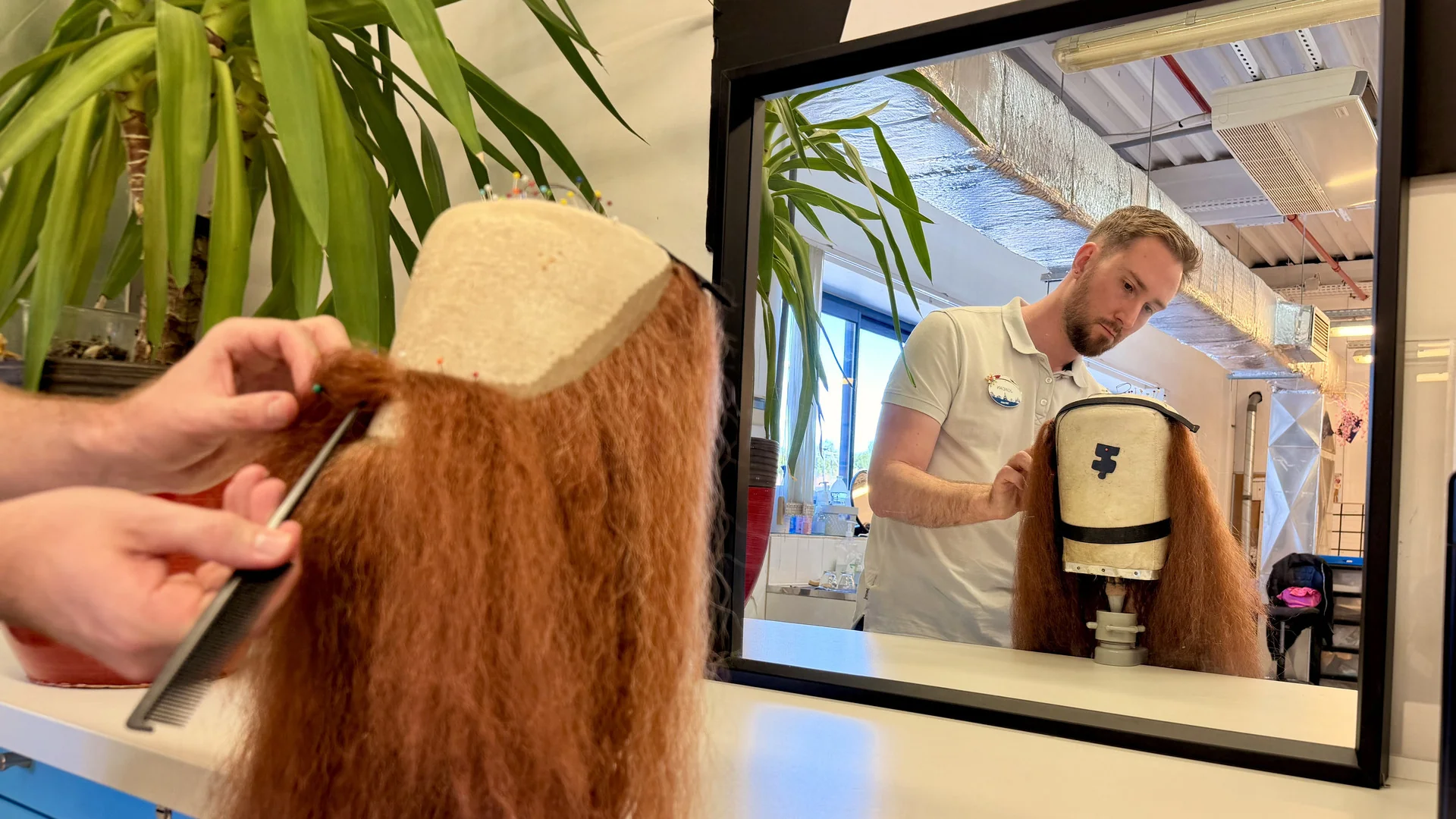 Dans une pièce bien éclairée agrémentée de plantes, une personne arrange une perruque rouge sur une tête de mannequin devant un miroir, donnant vie aux looks éclatants des princesses Disney grâce à un maquillage professionnel et à l'inspiration des créateurs de costumes.