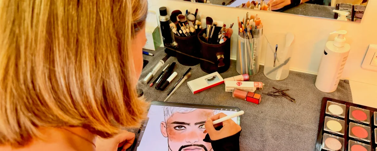 Personne utilisant une tablette numérique pour dessiner le visage d'un homme, entourée d'accessoires de maquillage, de perruques et de produits cosmétiques posés sur une coiffeuse munie d'un miroir.
