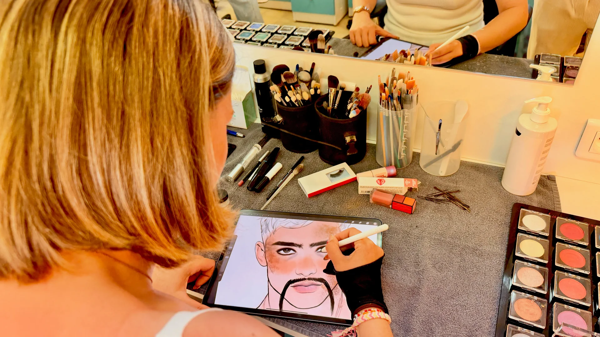 Personne utilisant une tablette numérique pour dessiner le visage d'un homme, entourée d'accessoires de maquillage, de perruques et de produits cosmétiques posés sur une coiffeuse munie d'un miroir.