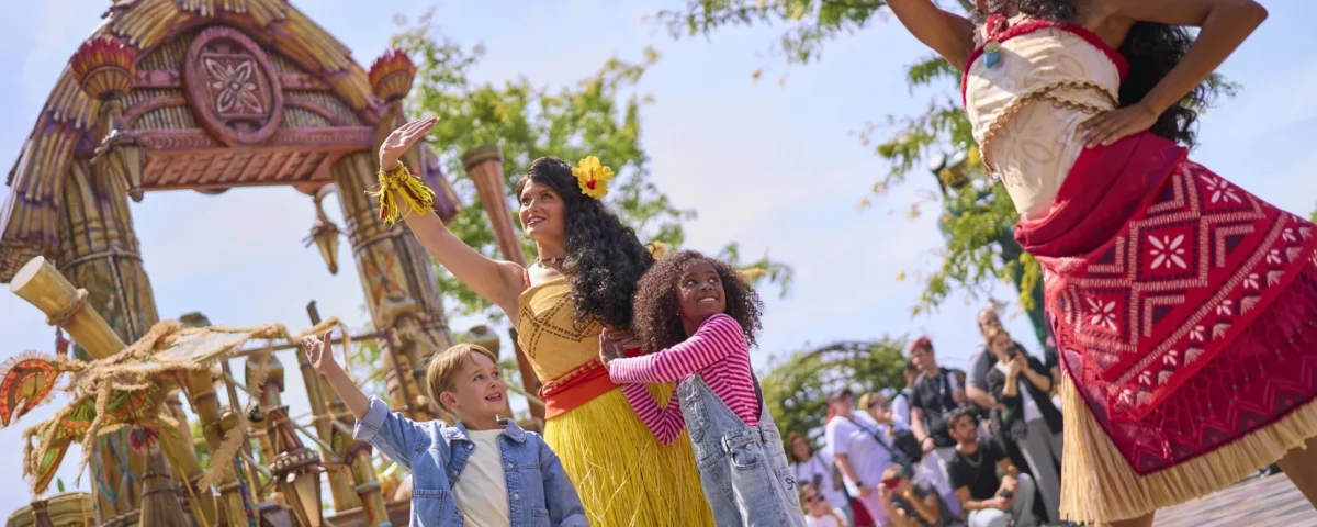Deux artistes vêtus de costumes polynésiens et deux enfants saluent la foule et dansent en plein air près d'une structure de style tiki, divertissant les spectateurs dans le cadre de la très animée « Cavalcade des Princesses Disney » à Disney Adventure World.