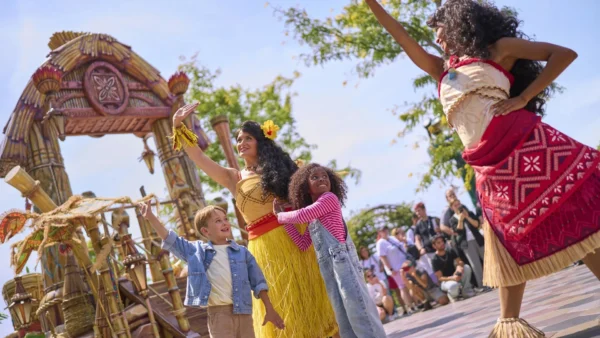 Deux artistes vêtus de costumes polynésiens et deux enfants saluent la foule et dansent en plein air près d'une structure de style tiki, divertissant les spectateurs dans le cadre de la très animée « Cavalcade des Princesses Disney » à Disney Adventure World.