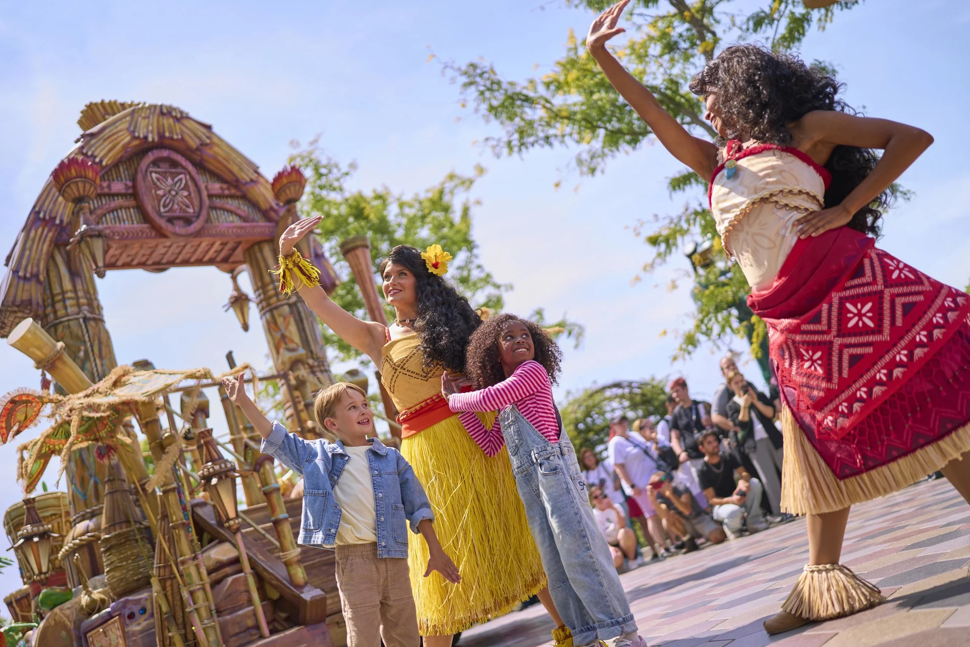 Deux artistes vêtus de costumes polynésiens et deux enfants saluent la foule et dansent en plein air près d'une structure de style tiki, divertissant les spectateurs dans le cadre de la très animée « Cavalcade des Princesses Disney » à Disney Adventure World.