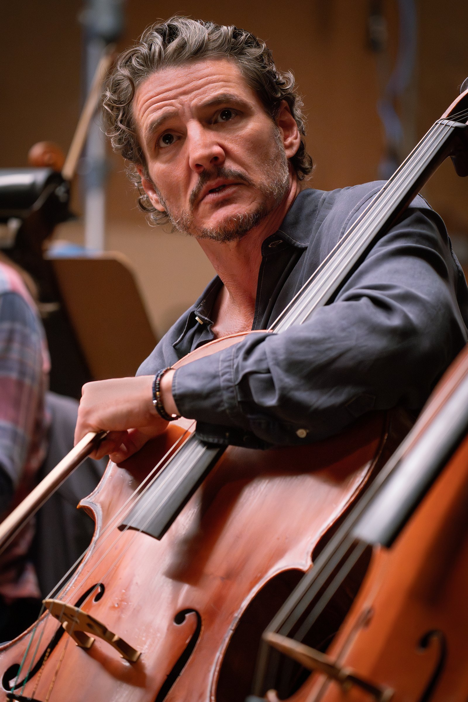 Un homme aux cheveux ondulés et barbu joue du violoncelle dans un orchestre ; vêtu d'une chemise sombre, il regarde légèrement sur le côté, l'air concentré — une silhouette saisissante qui rappelle Pedro Pascal jouant avec une intensité discrète, comme s'il incarnait la gravité cinématographique d'un drame de Tony Gilroy.