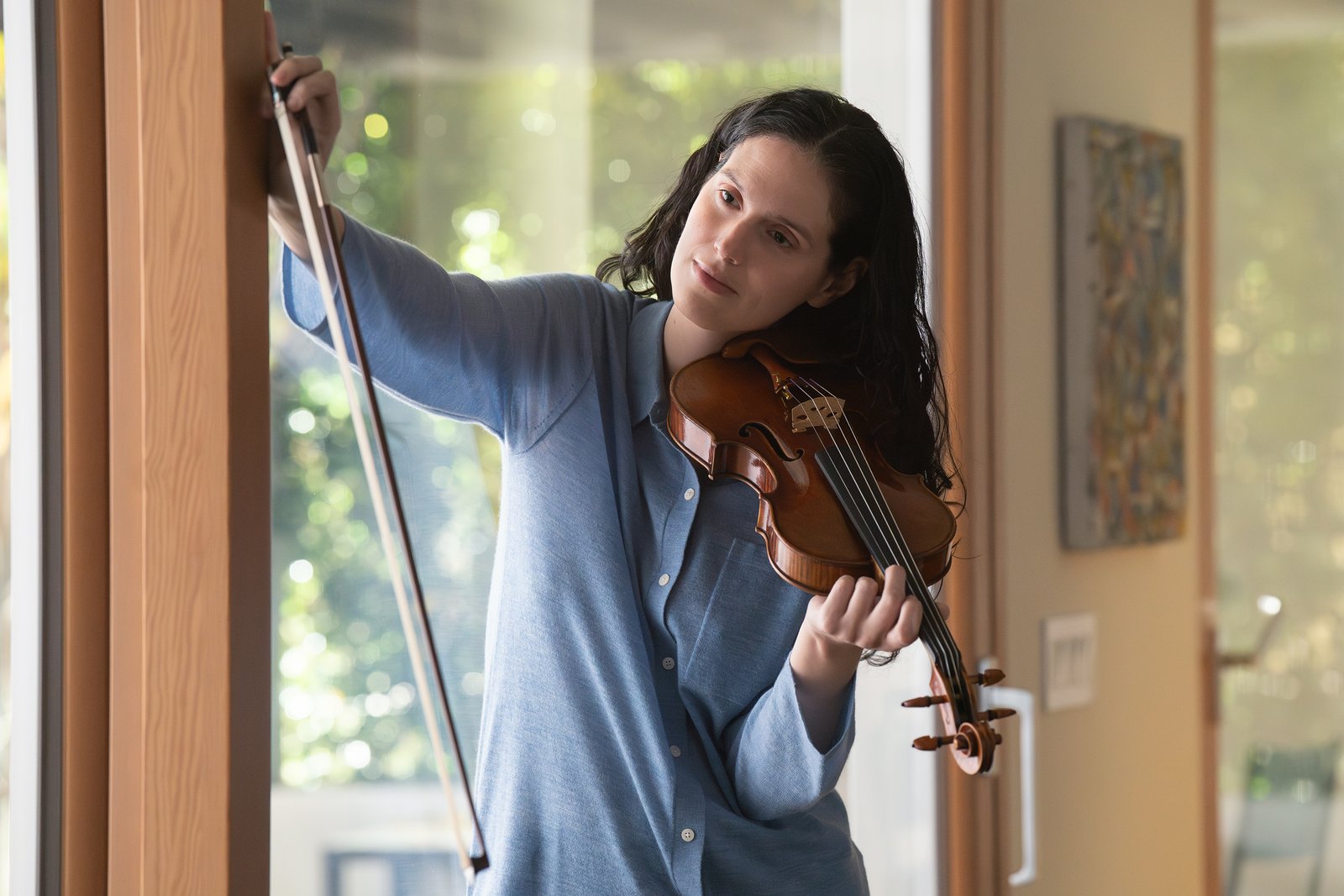 Une femme vêtue d'une chemise bleue joue du violon à l'intérieur, tenant l'archet d'une main et le violon de l'autre, tandis que la lumière du soleil pénètre à flots par une fenêtre derrière elle — une scène empreinte d'une intensité sereine qui rappelle la narration cinématographique de Tony Gilroy.