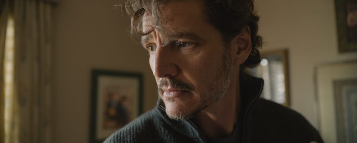 Un homme aux cheveux ondulés et à la barbe soigneusement taillée regarde sur le côté dans une pièce baignée d'une lumière tamisée, avec des photos encadrées accrochées au mur derrière lui, ce qui rappelle Pedro Pascal dans une scène pleine d'ambiance tirée d'une production de Tony Gilroy.