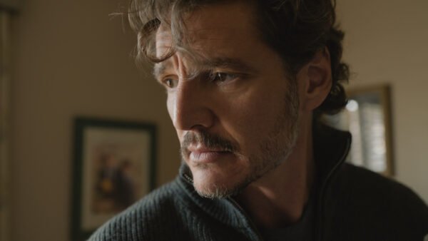 Un homme aux cheveux ondulés et à la barbe soigneusement taillée regarde sur le côté dans une pièce baignée d'une lumière tamisée, avec des photos encadrées accrochées au mur derrière lui, ce qui rappelle Pedro Pascal dans une scène pleine d'ambiance tirée d'une production de Tony Gilroy.