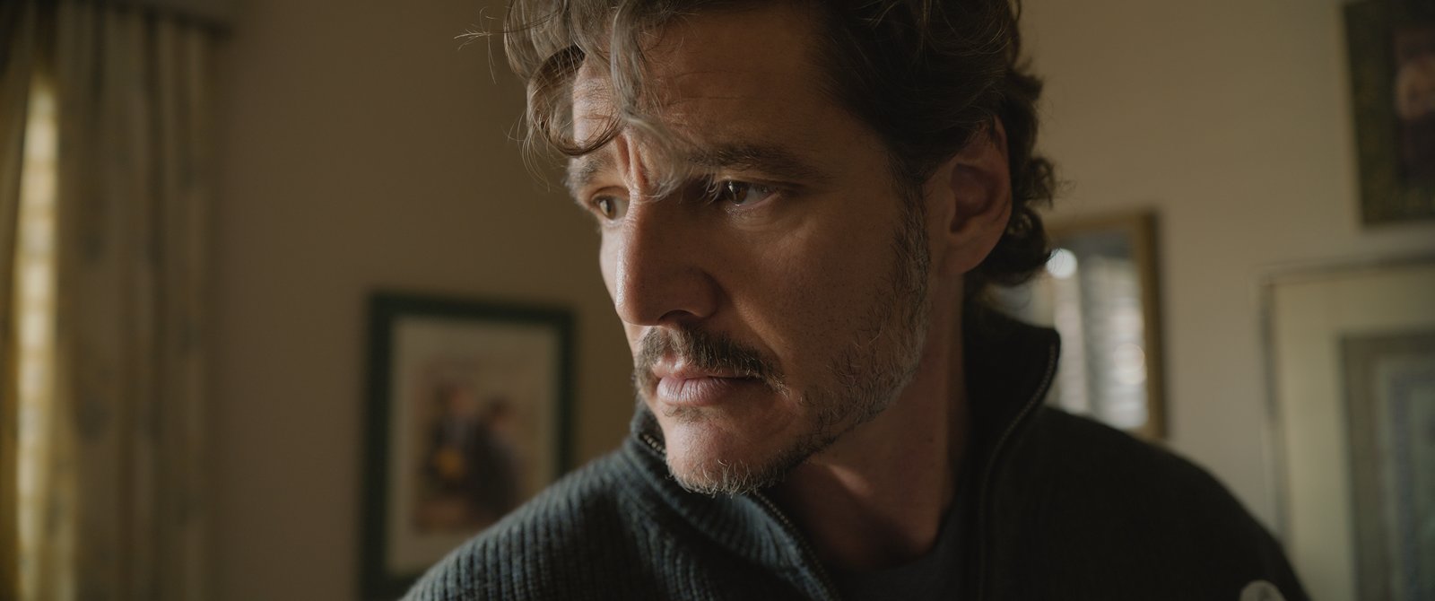 Un homme aux cheveux ondulés et à la barbe soigneusement taillée regarde sur le côté dans une pièce baignée d'une lumière tamisée, avec des photos encadrées accrochées au mur derrière lui, ce qui rappelle Pedro Pascal dans une scène pleine d'ambiance tirée d'une production de Tony Gilroy.