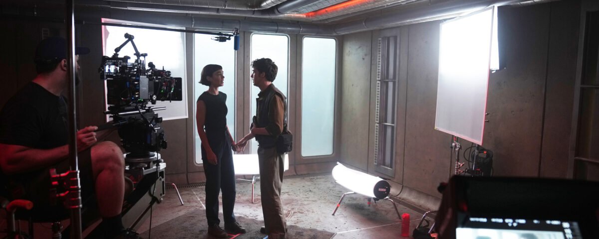 Deux acteurs se tiennent face à face sur un plateau de tournage, entourés de caméras, de l'équipe technique et de matériel d'éclairage, dans une salle au style industriel. L'énergie intense qui se dégage de la scène laisse supposer qu'il s'agit du tournage de la saison 2 d'Alien: Earth, avec Peter Dinklage qui attire tous les regards au centre de la scène.