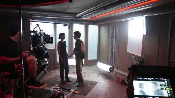 Deux acteurs se tiennent face à face sur un plateau de tournage, entourés de caméras, de l'équipe technique et de matériel d'éclairage, dans une salle au style industriel. L'énergie intense qui se dégage de la scène laisse supposer qu'il s'agit du tournage de la saison 2 d'Alien: Earth, avec Peter Dinklage qui attire tous les regards au centre de la scène.