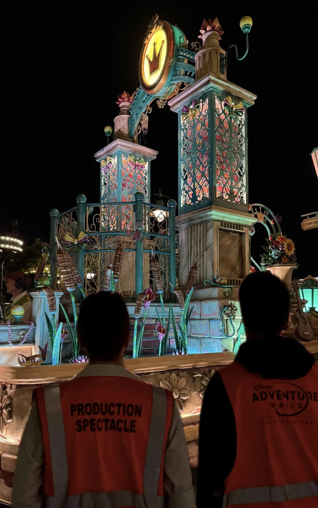 Deux personnes vêtues de gilets de sécurité se tiennent devant un char de parade richement décoré et brillamment éclairé, de nuit, orné d'une couronne et de décorations raffinées inspirées des princesses Disney. Derrière elles, ce spectacle éblouissant témoigne du talent artistique des créateurs de costumes et des maquilleurs experts, qui donnent vie à des contes de fées tant appréciés.