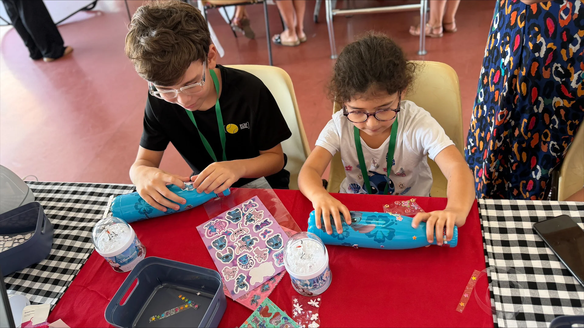 Deux enfants sont assis à une table et décorent des objets bleus avec des autocollants de Mickey et Minnie. Du matériel de loisirs créatifs et des feuilles d'autocollants sont disposés devant eux sur une nappe rouge, apportant une touche de la magie de Disneyland Paris à leur après-midi créatif.
