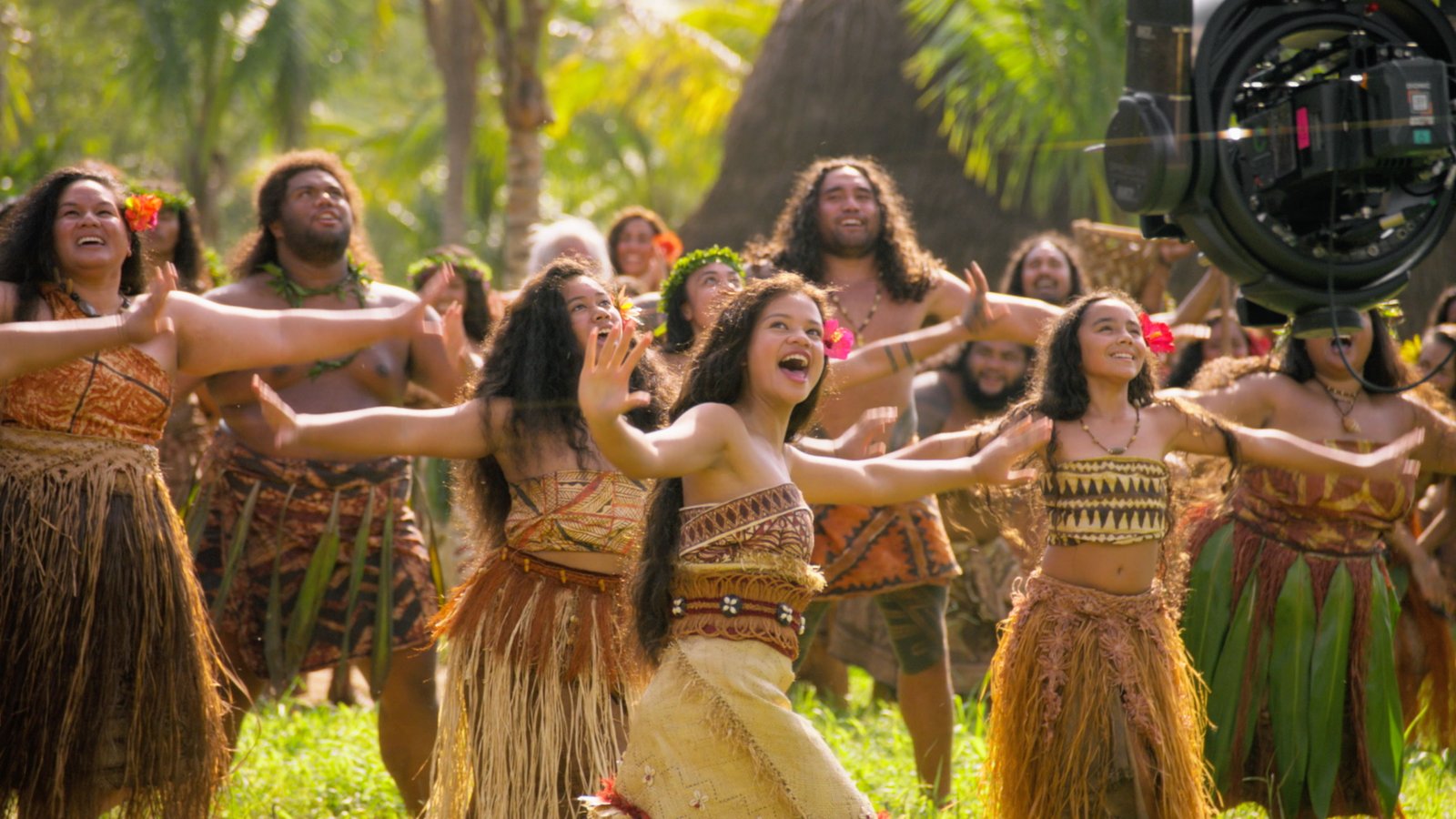 Un groupe de personnes vêtues de costumes traditionnels polynésiens dansent et chantent en plein air, évoquant l'esprit du film « Vaiana » de Disney ; une caméra de cinéma, visible à droite de l'image, met en valeur cette scène pleine de vie qui rappelle la magie du cinéma.