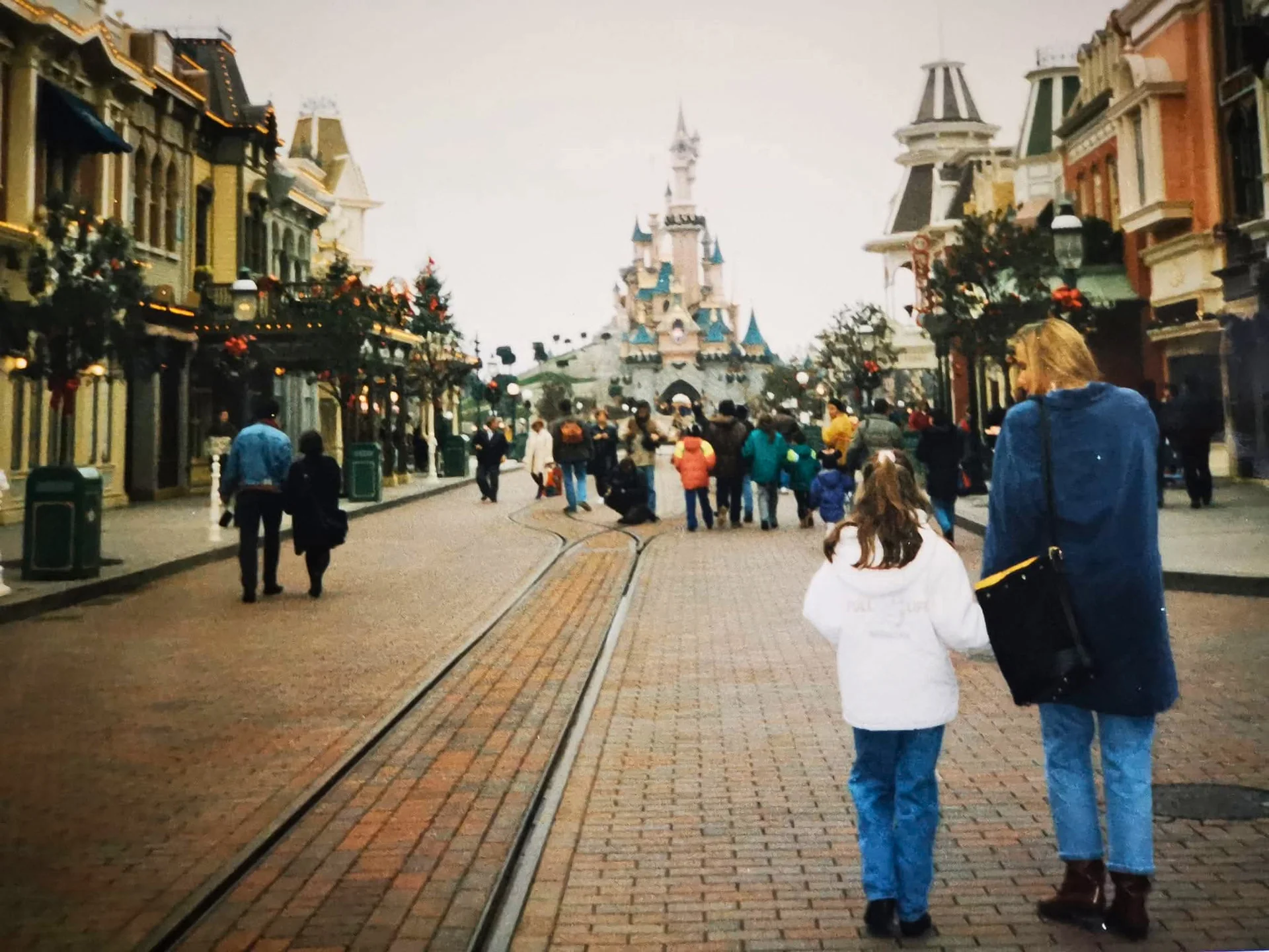 Des visiteurs marchent le long d’une rue pavée en direction d’un château à Disneyland Paris, bordée de boutiques de part et d’autre et traversée par des rails au milieu — 34 ans plus tard, c’est comme un vœu exaucé pour ceux qui rêvaient de magie depuis leur enfance.