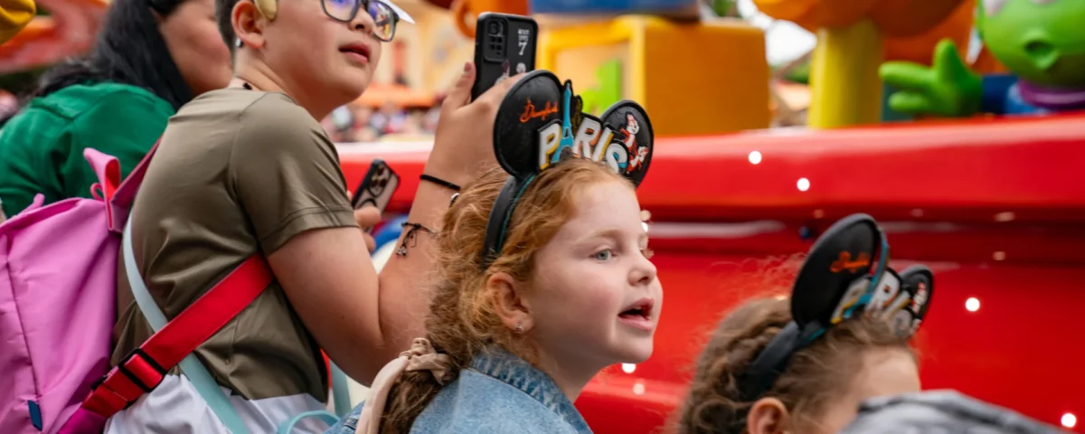 Des enfants portant des oreilles de Mickey Mouse de Disneyland Paris regardent un char coloré défiler. L'un d'eux tient un appareil photo, tandis qu'un autre porte un bandeau vert à l'effigie d'un personnage. Des décorations aux couleurs vives sont visibles à l'arrière-plan. Ils immortalisent ces moments inoubliables avant de se rendre au bureau des objets non réclamés de Disneyland Paris pour récupérer d'éventuels objets perdus.