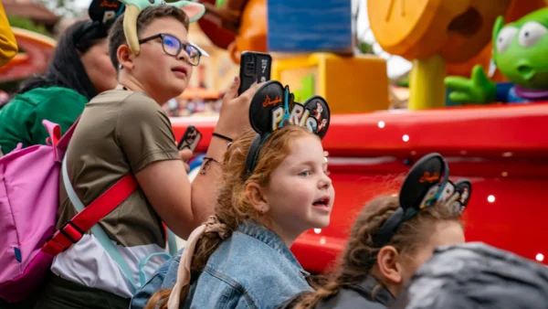 Des enfants portant des oreilles de Mickey Mouse de Disneyland Paris regardent un char coloré défiler. L'un d'eux tient un appareil photo, tandis qu'un autre porte un bandeau vert à l'effigie d'un personnage. Des décorations aux couleurs vives sont visibles à l'arrière-plan. Ils immortalisent ces moments inoubliables avant de se rendre au bureau des objets non réclamés de Disneyland Paris pour récupérer d'éventuels objets perdus.