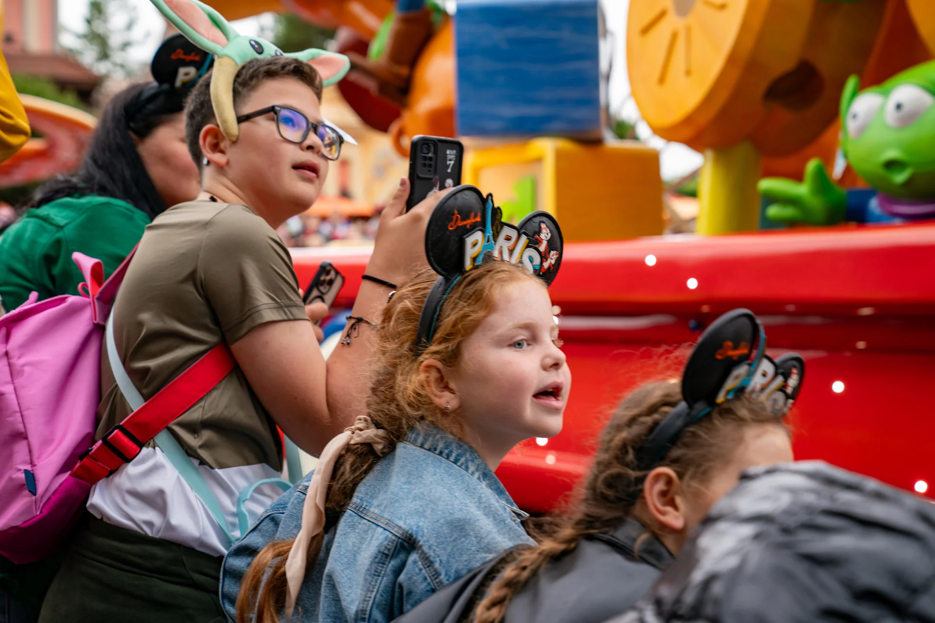 Des enfants portant des oreilles de Mickey Mouse de Disneyland Paris regardent un char coloré défiler. L'un d'eux tient un appareil photo, tandis qu'un autre porte un bandeau vert à l'effigie d'un personnage. Des décorations aux couleurs vives sont visibles à l'arrière-plan. Ils immortalisent ces moments inoubliables avant de se rendre au bureau des objets non réclamés de Disneyland Paris pour récupérer d'éventuels objets perdus.