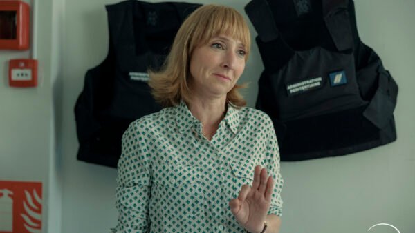Une femme aux cheveux châtain clair se tient à l'intérieur, faisant un geste de la main droite. Deux gilets pare-balles noirs sont accrochés au mur derrière elle, créant une atmosphère tendue qui rappelle un environnement de surveillance. Le logo Disney+ est visible dans un coin, laissant entrevoir une nouvelle série Disney+ dont le lancement est prévu le 16 septembre.