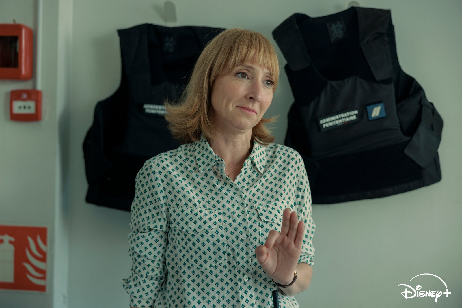 Une femme aux cheveux châtain clair se tient à l'intérieur, faisant un geste de la main droite. Deux gilets pare-balles noirs sont accrochés au mur derrière elle, créant une atmosphère tendue qui rappelle un environnement de surveillance. Le logo Disney+ est visible dans un coin, laissant entrevoir une nouvelle série Disney+ dont le lancement est prévu le 16 septembre.