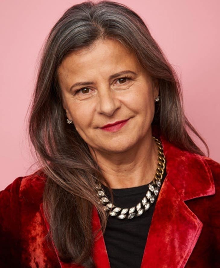 Une femme aux longs cheveux gris, vêtue d'un blazer en velours rouge, d'un haut noir et d'un collier en argent, se tenant debout devant un fond rose : son style assuré s'intégrerait parfaitement dans « Alien : Earth », saison 2, ou même dans un sketch de Tracey Ullman.