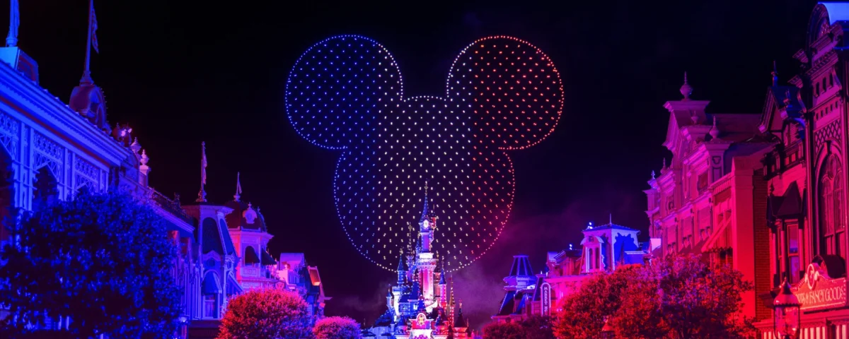 Un « Spectacle Nocturne » éblouissant à Disneyland Paris projette une immense silhouette représentant la tête de Mickey Mouse au-dessus du château, tandis que les bâtiments sont illuminés de bleu et de rouge et qu'une foule s'est rassemblée en contrebas pour célébrer le 14 juillet.