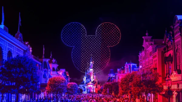 Un « Spectacle Nocturne » éblouissant à Disneyland Paris projette une immense silhouette représentant la tête de Mickey Mouse au-dessus du château, tandis que les bâtiments sont illuminés de bleu et de rouge et qu'une foule s'est rassemblée en contrebas pour célébrer le 14 juillet.